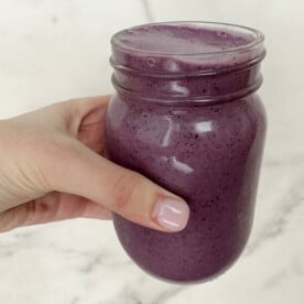 A hand holding a blueberry banana spinach smoothie in a clear jar