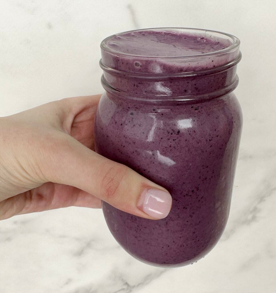 A hand holding a blueberry banana spinach smoothie in a clear jar