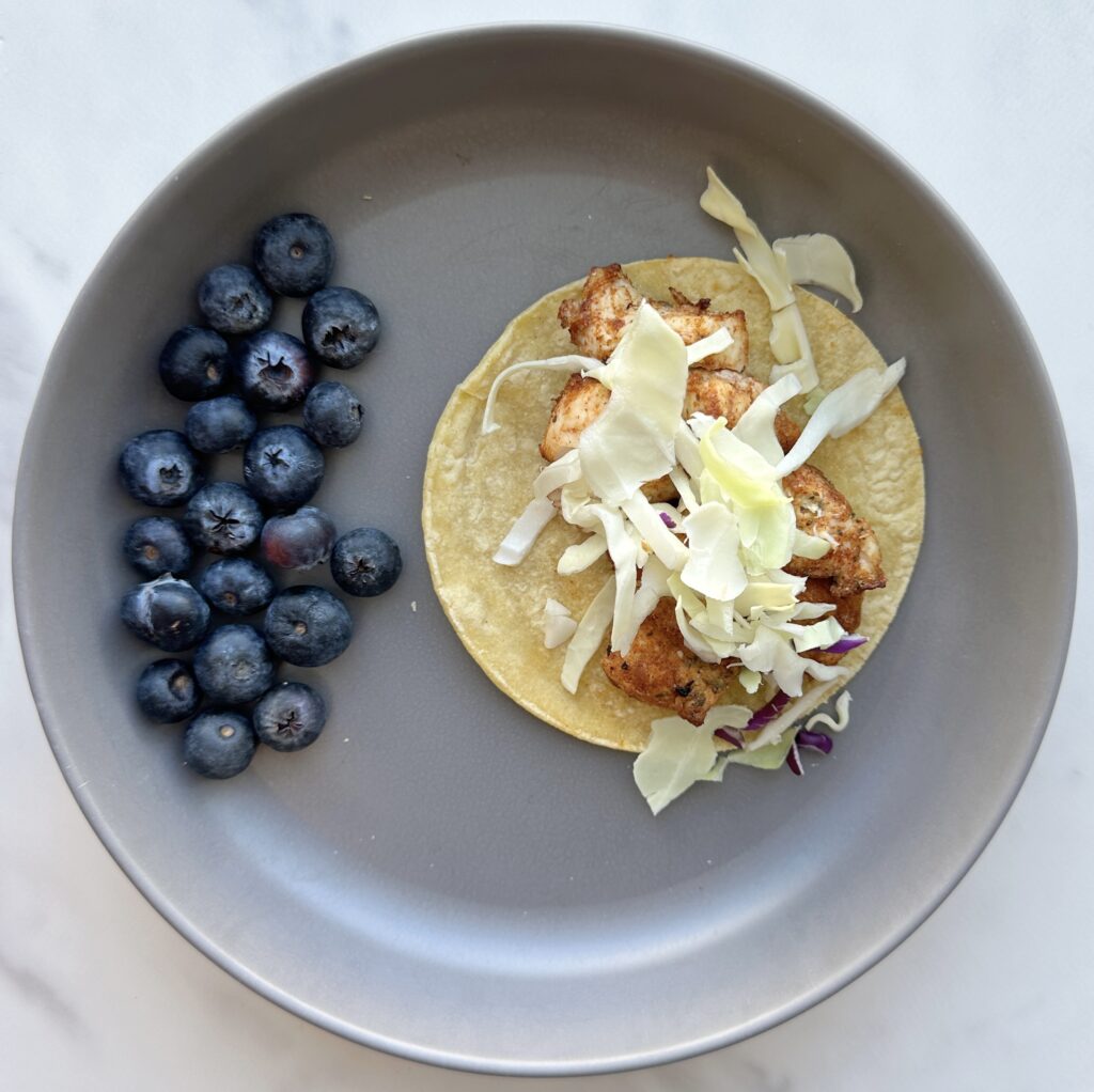 A small crispy chicken taco topped with lettuce on a gray plate beside several blueberries.