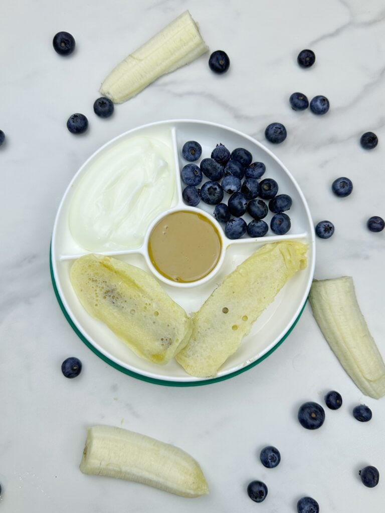 Banana dippers on a white divided plate with peanut butter, blueberries, and whipped cream