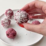 A hand holding up a raspberry bliss ball in front of a white plate with several raspberry bliss balls.